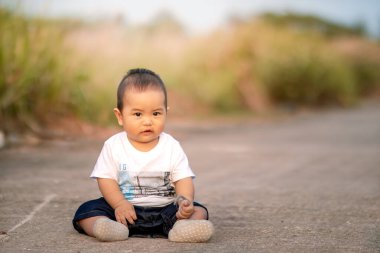 Yolda oturan sevimli Asyalı bebek. Bebek, kameraya doğru tatlı tatlı gülümserken yabani otları tutuyor. Öğleden sonra fotoğraf atmosferi.
