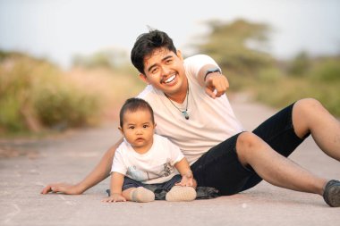 Baba ve bebek kamerayı işaret ederken oturuyorlar. Beyaz çift kıyafeti giyiyorlardı. Açık havada güneşli bir öğleden sonra fotoğraf atmosferi.