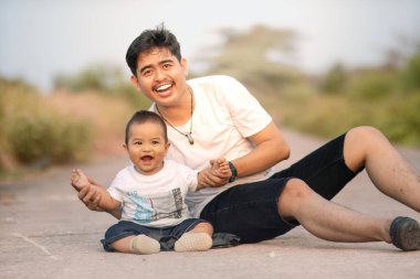 Baba ve oğul otururken, baba oğlana sataşıyor ve neşeyle gülüyor. Beyaz çift kıyafeti giyiyorlardı. Açık havada güneşli bir öğleden sonra fotoğraf atmosferi.
