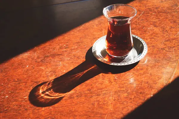 A glass of Turkish tea on the wooden table with the sun shining on it - special Turkish tea glass and decorated small metal plate - tradition of Turkey
