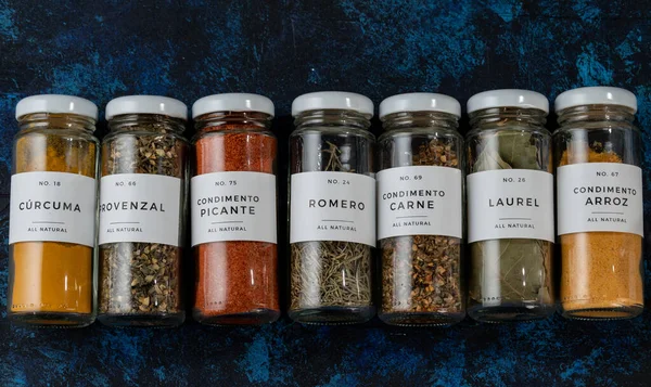 stock image row of several glass jars, lying on a blue table. the jars are elongated. they have different seasonings, such as spicy, bay leaf, turmeric, seasoning for meat and for rice, provenzal.