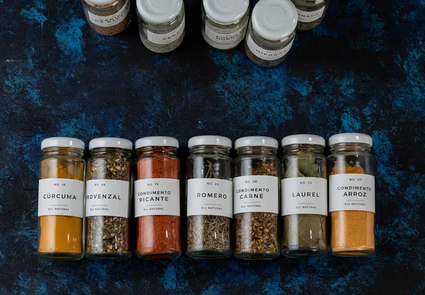row of several glass jars, lying on a blue table. the jars are elongated and have a white lid. they have different seasonings, such as spicy, bay leaf, turmeric, seasoning for meat and for rice.