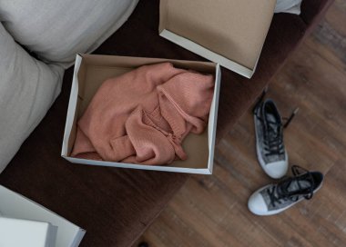 Kahverengi kanepede açık beyaz bir hediye kutusunun üst görüntüsü. Kutuda pembe bir kazak var. Diğer beyaz pullar bir tarafa istiflenmiş olarak görülebilir. Bir çift de ayakkabı var.