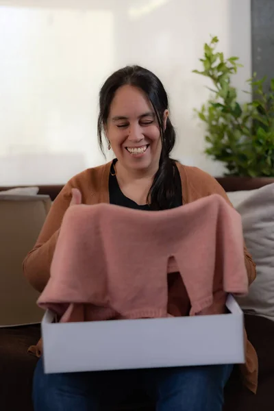 a happy and smiling lady is opening a white gift box. looking at a pink sweater that came inside the box. She is sitting in an armchair in the living room of the house. The sweater is light pink.