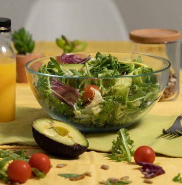 Masanın üzerinde bir kase dolusu cam içinde salata. Domates, marul, turp ve avokado var. Meyve suyu şişesi de var. Domatesler kirazlı. Arkadan bakıldığında, orada geniş yapraklı otlar görürsün..