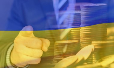 Bu resim, Ukrayna bayrağının renkleriyle örtülü, yönü ve odaklanmayı sembolize eden, ileri bakan bir elin yakın çekimini gösteriyor. Arka planda madeni para yığınları mali büyüme, yatırım ve ekonomik istikrarı temsil ediyor. Kombina.