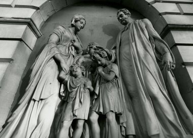 A detailed stone sculpture of two adults and three children in classical attire, set within an arched niche, displaying intricate craftsmanship and evoking a scene of familial or educational significance clipart