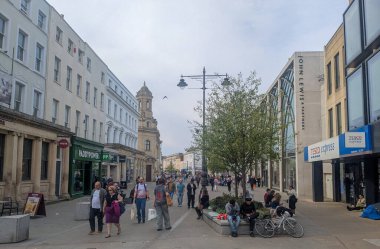 Cheltenham, İngiltere - 12 Nisan 2025: Alışverişçiler bulutlu bir günde John Lewis ve Tesco Express gibi dükkanlarla dolu yaya geçidi boyunca yürüyorlar