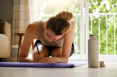 Genç beyaz kadın, evde yoga minderinin üzerinde minderin yanında duran su şişesiyle günlük tutuyor. Sağlık ve Sıhhat kavramı.