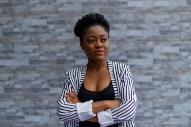 Portrait of confident young female professional with afro hair and arms crossed standing against brick wall outside office