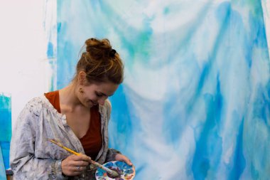 Stüdyodaki kadın ressamın geniş portresi arka planda büyük bir tuvalle bir palette boya karıştırıyor.