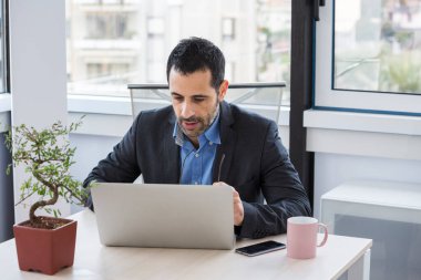 Ceket ve gömlek giymiş bir yönetici çalışma odasında oturuyor.