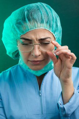 Laboratuvar önlüğü, kep ve cerrahi maske takmış gözlüklü kadın doktor yeşil arka planda izole edilmiş hastaya tepeden bakıyor.
