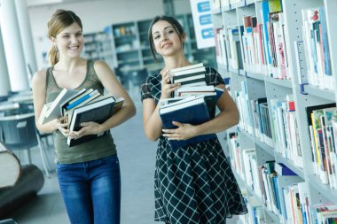Kütüphanede elinde kitaplarla duran iki genç kız öğrenci.