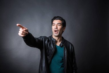 Koyu renk saçlı, yeşil tişörtlü, deri ceketli koyu renk arka planda sırıtan bir adam.