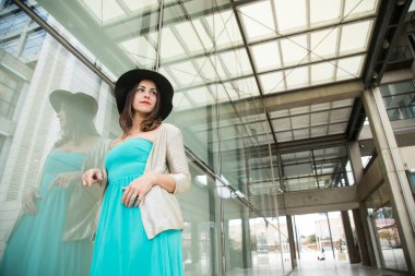 Zümrüt yeşili elbiseli esmer kız ve şapkası modern bir şehir ortamında duvara yaslanırken ayakta duruyor.