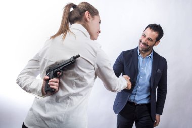 Beyaz gömlekli sarışın kız elinde silah tutarken siyah saçlı bir adamla el sıkışıyor. Ceket ve gömlek giymiş, beyaz arka planda soyutlanmış.