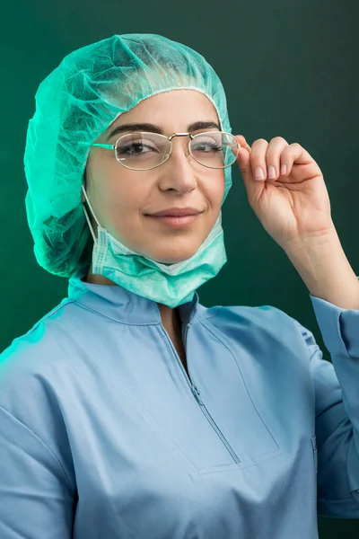身穿实验室外套 头戴帽子 戴着外科口罩的女医生看上去很严肃 她的眼镜被绿色背景隔开了 — 图库照片