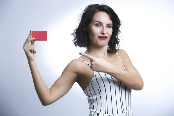 White woman with black hair dressed in a white striped jumpsuit points to a red card, isolated on white background