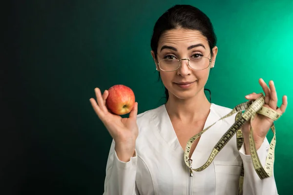 白衣の栄養士は 人間の体を測定するためのメートルと誇らしげに示すリンゴを手に持っています 緑の背景に隔離された — ストック写真
