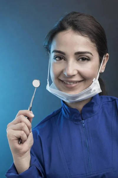 Female dentist with surgical mask and blue coat shows work tools, isolated on light blue background