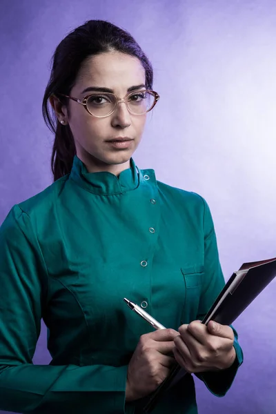 身穿绿色实验室外套的女医生 头戴黑色文件夹和眼镜 眼神严肃 与紫色背景隔离 — 图库照片