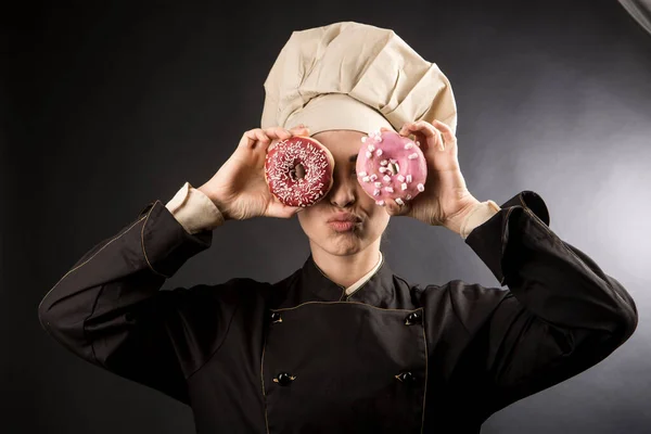 微笑的厨师很好地运用彩色甜甜圈 把它们当作望远镜 用黑色背景隔开 — 图库照片
