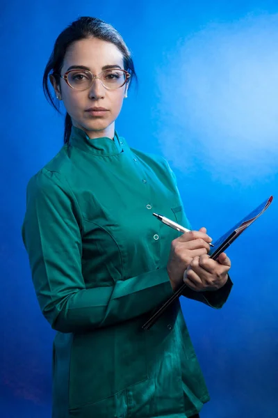 報告書と緑の研究室のコートの眼鏡とフォルダの美しい女性医師 青の背景に隔離された — ストック写真