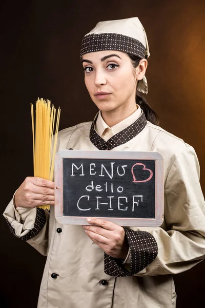 女性料理人の黒板に シェフのメニュー と書かれ 生のスパゲティを手に持ち 茶色の背景に隔離された — ストック写真
