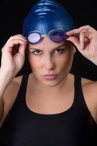 黒の水着を身に着けている水泳の肖像画彼女のゴーグルを脱ごうとしていると黒の背景に隔離された決定的かつ集中的な外観 青の帽子で見えます — ストック写真