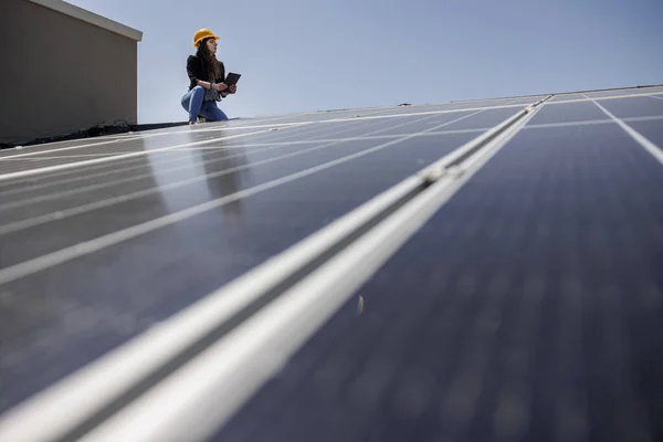 Female engineer in a yellow hard hat checks the condition of a photovoltaic system on the roof of a house with a tablet in her hand