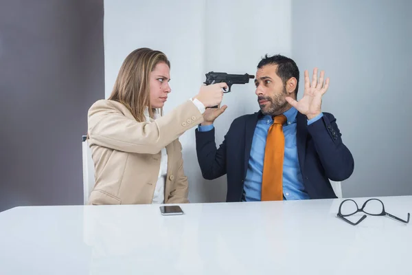 两个同事坐在办公室里 秘书生气地用枪指着她吓坏了的老板 — 图库照片