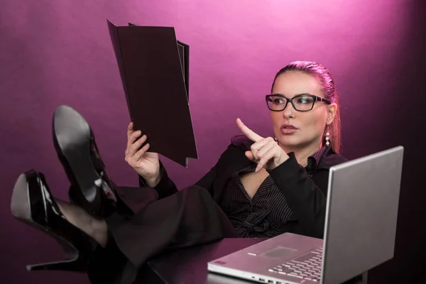 眼鏡をかけた回収髪を持つ女性マネージャー テーブルの上に足でコンピュータ上で作業黒のビジネススーツを身に着けています 赤の背景に隔離されました — ストック写真
