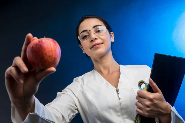穿着实验室外套的营养师 她的头发收集在一只手拿着一个苹果和一个带尺寸的带子 用蓝色的背景隔开 — 图库照片