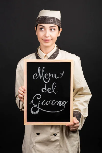 制服姿の笑顔のシェフが イタリア語で メニュー ジョルノ と書かれた黒板を手に持っており オレンジを背景に孤立した 当時のメニュー を意味する — ストック写真