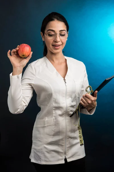 身穿白衣的营养师手里拿着一个测量人体的仪器 还有一个苹果 它骄傲地在绿色的背景下显得孤立无援 — 图库照片