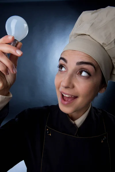 穿着厨师制服的女厨师手里拿着一个灯泡 表示她正在寻找一个创意配方的好主意 这个配方被淡淡的背景隔开了 — 图库照片