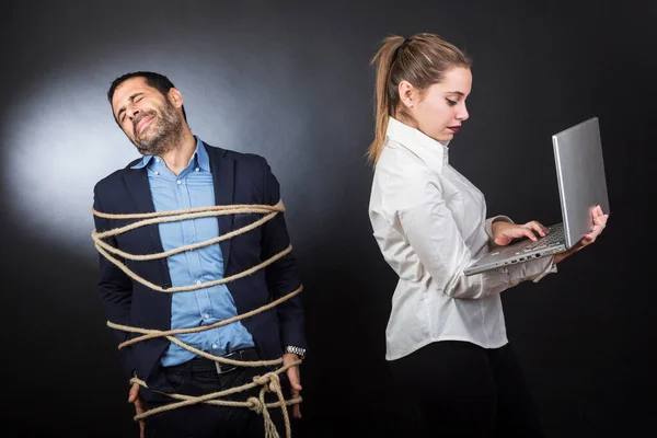 man in jacket tied with a rope is desperate in front of a blonde girl who controls the laptop, isolated on black background.