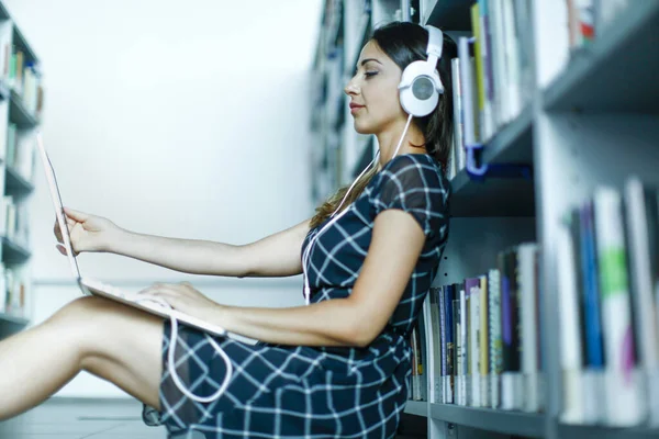一个带着耳机的年轻的黑发女孩坐在图书馆的架子上 在电脑上工作 — 图库照片
