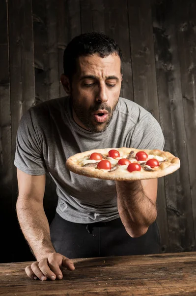 一个留着胡子的黑头发男人正要吃一个带有樱桃西红柿的披萨 它是木制背景的 — 图库照片