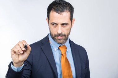 Koyu saçlı, sakallı, gömlekli, turuncu ceketli ve kravatlı bir adam, beyaz arka planda izole edilmiş, varsayımsal saydam bir yüzeye işaret çiziyor.