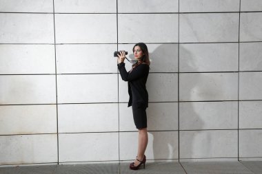 Siyah takım elbiseli esmer kadın yönetici, elindeki dürbünle diğer tarafa bakıyor, beyaz dikdörtgen duvar arka planında izole edilmiş.