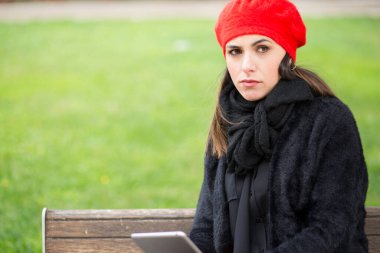 Siyah giyimli, kırmızı kulaklıklı bir kız parkta bankta oturuyor.