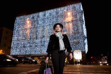 Kahverengi saçlı, paltolu ve siyah şapkalı güzel bir kadın alışverişe gitmek için dükkanlarda dolaşıyor.