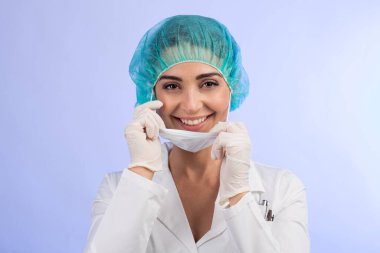 Beyaz önlüklü gülümseyen kadın doktor maskeyi tutuyor ve doğrudan kameraya bakıyor.