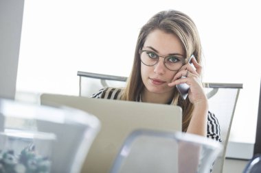 Çizgili gömlekli genç sarışın ofis çalışanı çalışma yerinde oturmuş bilgisayarının önünde telefonla konuşuyor.