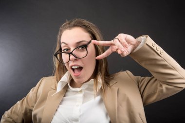 Yüzünde bej ceketli, beyaz gömlekli, çarpık gözlüklü sarışın kız parmaklarıyla zafer işareti yapıyor.