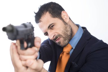 Koyu saçlı, sakallı, turuncu ceketli ve kravatlı adam boş bir silahı işaret ediyor. 