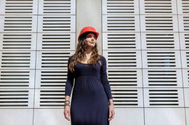 Donna vestita elegante con elmetto di protezione rosso in posizione statica guarda seria in lontananza in contesto urbano moderno
