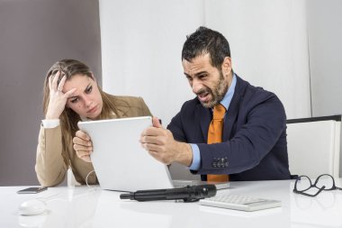 Koyu renk saçlı, sakallı, turuncu takım elbiseli, kravatlı, ofisinde umutsuzluğa kapılmış, yanında sekreteriyle bilgisayarının önünde oturuyor.